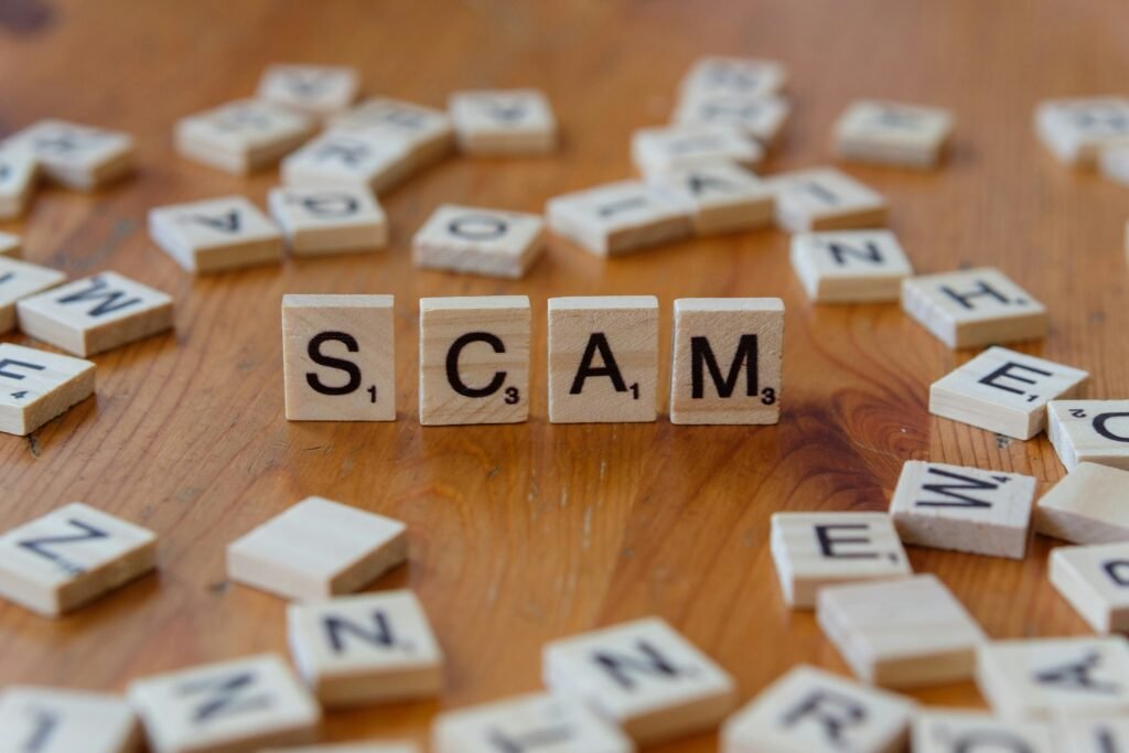 Wooden letter tiles spelling “SCAM” on a table, surrounded by scattered letters, symbolizing confusion and hidden online fraud.