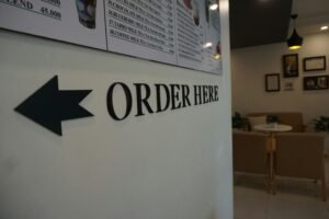 A clean white background with bold black lettering spelling "Order Here" in a simple, straightforward font, resembling a directional sign or store notice.